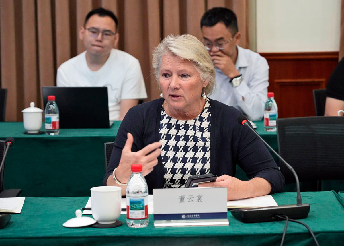 Susan Thornton, a woman with blond hair and fair skin, is seated at a conference table and is speaking into a microphone. Two men, both with short dark hair and glasses, are seated behind her, listening to her remarks.